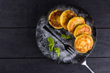 Cottage cheese pancakes on a dark background. Syrniki with fresh mint. Pancakes with cottage cheese on a black plate sprinkled with powdered sugar. Healthy food