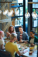 Group of young friendly people in casualwear sitting by served table at dinner and chatting