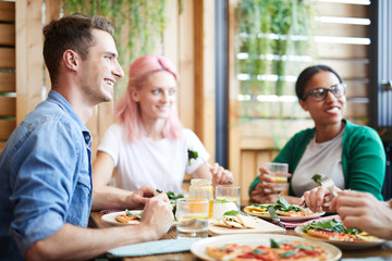 Three happy young friends sitting by served table and looking at their buddy during lunch