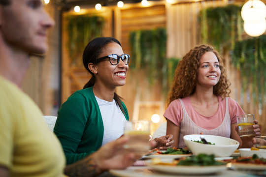 Two happy intercultural girls sitting by served festive table at dinner and talking