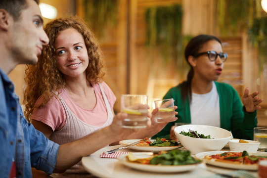 Smiling girl with wavy hair looking at her boyfriend during conversation by served table at dinner with their friends