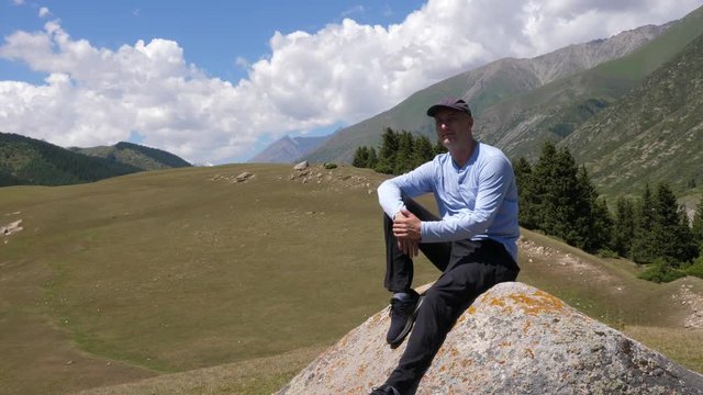 Handsome man sitting on large stone on mountain valley and green hills landscape