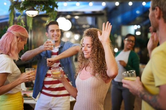 Excited young woman with drink dancing among group of friends and enjoying party