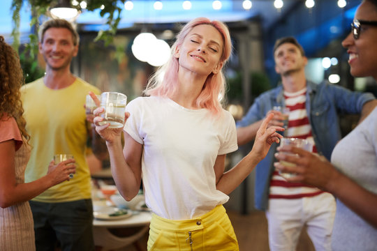 Excited girl in casualwear holding drink and dancing on background of her friends