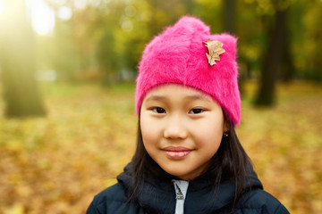 Cute girl with dry oak leaf on her pink beanie looking at you during chill on weekend