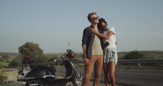 A Latino girl and a young blonde boy are standing on the road near their retro motorbike and are looking in the distance