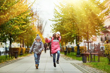 Two funny kids in warm casualwear jumping over trottoire on their way to school on October morning