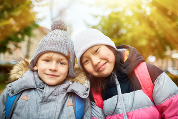 Cute intercultural kids in warm jackets and beanies looking at you in urban environment