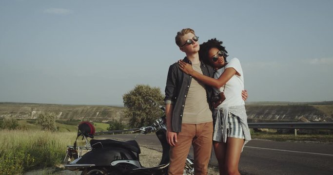 A beautiful couple wearing sunglasses are sitting by the road near their motorbike and are staring straight into the camera