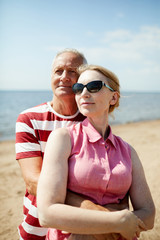 Aged man embracing his wife in sunglasses while both enjoying their rest by seaside on summer day