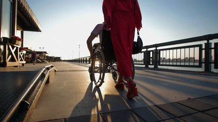 A girl with red hair in orange dress rolls a disabled guy in a wheelchair on the waterfront in the sunset