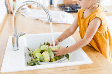 Cute little girl wearing linen mustard dress washing pears and apples in the sink in the white kitchen scandinavian style