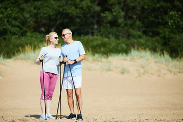 Cheerful senior active couple standing on sand and having talk while trekking on summer day