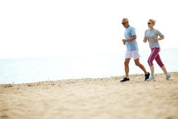 Aged man and woman in activewear jogging on sandy beach along seashore