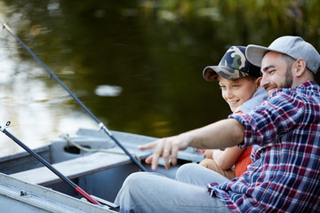 Cheerful father showing to his son a good place for fishing while they sitting in the boat