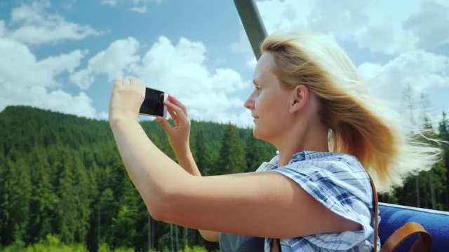 A middle-aged woman takes pictures of beautiful landscapes, sits on a bench of a ski lift. Holidays in the mountains in summer