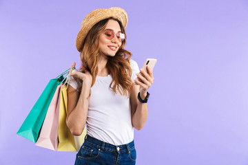 Happy cute woman isolated over purple wall background holding shopping bags using mobile phone.