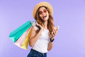 Happy cute woman isolated over purple wall background holding shopping bags using mobile phone.