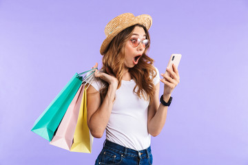 Screaming cute woman isolated over purple wall background holding shopping bags using mobile phone.