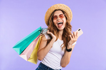 Screaming cute woman isolated over purple wall background holding shopping bags using mobile phone.