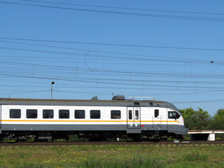 Fototapeta premium Electric train riding in the countryside among green grass and wildflowers. Railway locomotive against the blue sky and electric wires