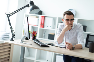 A young man in glasses stands near a table in the office and holds a pencil in his hand.