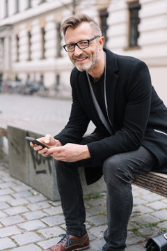 eleganter mann sitzt in der stadt auf einer bank und hält sein smartphone in der hand
