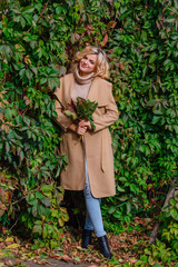 Young beautiful woman in sweater and coat stands next to the background of wild grapes holding bouquet of autumn leaves