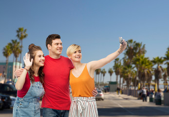 travel, tourism and summer holidays concept - group of happy smiling friends taking selfie by smartphone and hugging over venice beach background in california