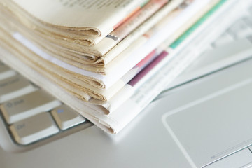 Newspapers and laptop. Pile of daily papers with news on the computer. Pages with headlines, articles folded and stacked on keypad of electronic device. Modern gadget and old journals, focus on paper 