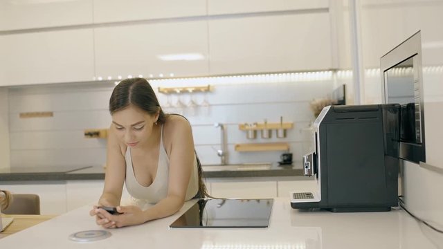 Beautiful woman typing message on her mobile phone at home. Young lady standing in the kitchen, near coffeemachine and oven and discussing plans for weekwnd with her friends using her smartphone