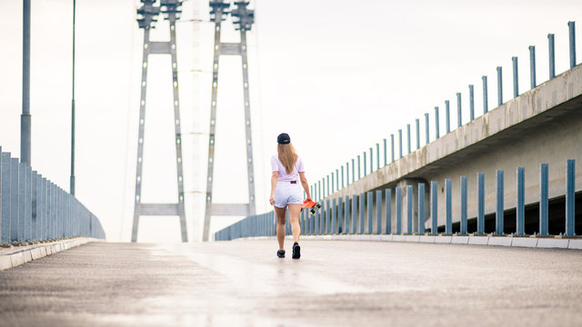 Young Blonde Girl Walking Away with Orange Skateboard on the Bridge