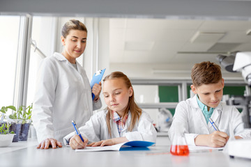 education, science and children concept - teacher and students studying chemistry at school laboratory and writing to workbooks