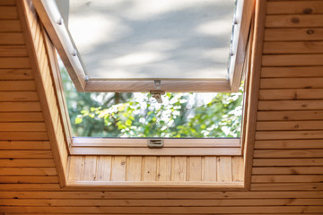 Opened roof window with blinds or curtain in wooden house attic. Room with slanted ceiling made of natural eco materials and park view through opened window. Environment friendly house