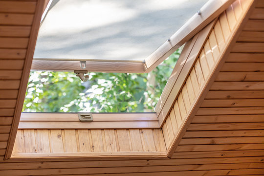 Fototapeta Opened roof window with blinds or curtain in wooden house attic. Room with slanted ceiling made of natural eco materials and park view through opened window. Environment friendly house