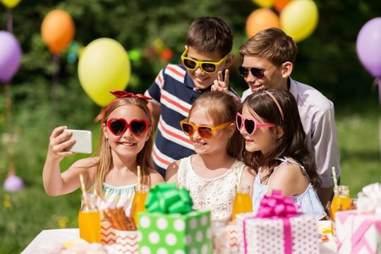holidays, childhood and technology concept - happy kids in sunglasses taking selfie by smartphone on birthday party at summer garden