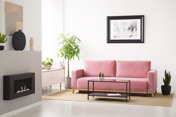 Poster above pink sofa between plants in white living room interior with fireplace. Real photo