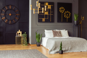 Real photo of a spacious bedroom interior with grey walls, clock, paintings, plants, bed and golden...