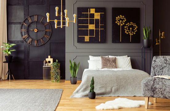 Real photo of golden accents, clock, paintings, plants and double bed in a dark bedroom interior