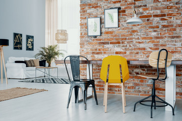 Chairs at dining table against red brick wall with posters in open space interior with white sofa. Real photo