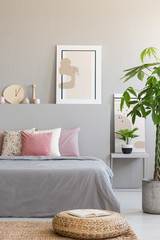 Pouf and plant next to grey bed with pink cushions in bedroom interior with poster. Real photo