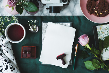 Top view of white paper sheets, coffee, rose, stamps, cake, camera on a green background
