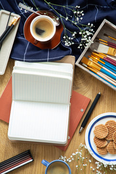 Messy desk with a notebook, coffee, pens, cookies and brushes. Top view