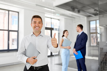 real estate business, sale and people concept - happy smiling realtor or businessman with folder showing thumbs up and customers at new office room