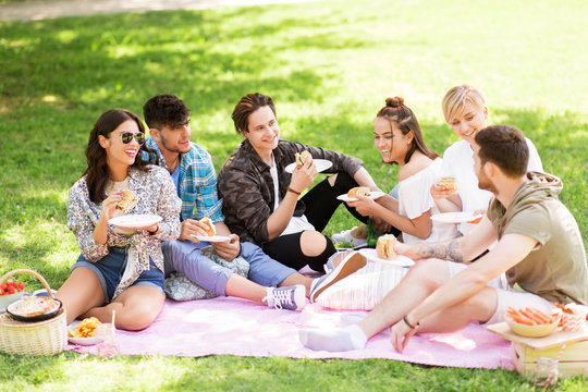 friendship, leisure and fast food concept - group of happy friends eating sandwiches or burgers at picnic in summer park