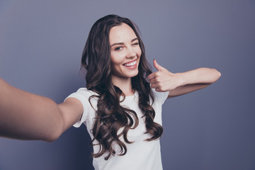 Self-portrait of magnificent lovely adorable gorgeous stylish nice confident curly-haired brunette girl in casual white t-shirt, showing thumb-up, winking, isolated over grey background