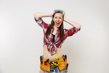 Strong young handyman woman in plaid top shirt, denim shorts, kit tools belt full of instruments in protective goggles isolated on white background. Female in male work. Copy space for advertisement.
