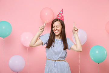 Portrait of overjoyed young woman with closed eyes in birthday hat and blue dress pointing index...