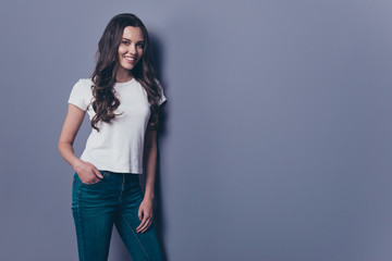 Charming magnificent attractive pretty lovely stylish nice cheerful dreamy curly-haired brunette girl in casual white t-shirt and jeans, holding hand in pocket, copy-space, isolated on grey background