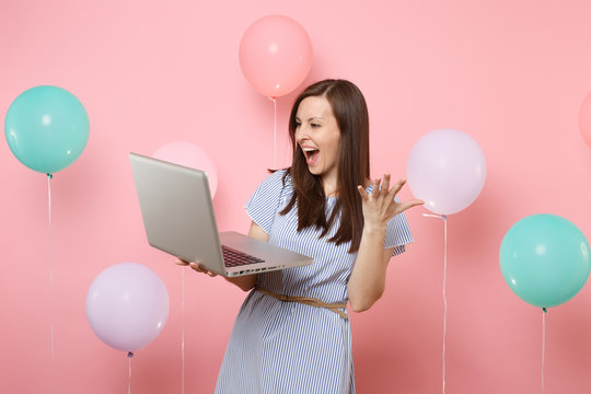 Portrait of overjoyed happy young woman in blue dress holding using laptop pc computer spreading hands on pink background with colorful air balloons. Birthday holiday party people sincere emotions.